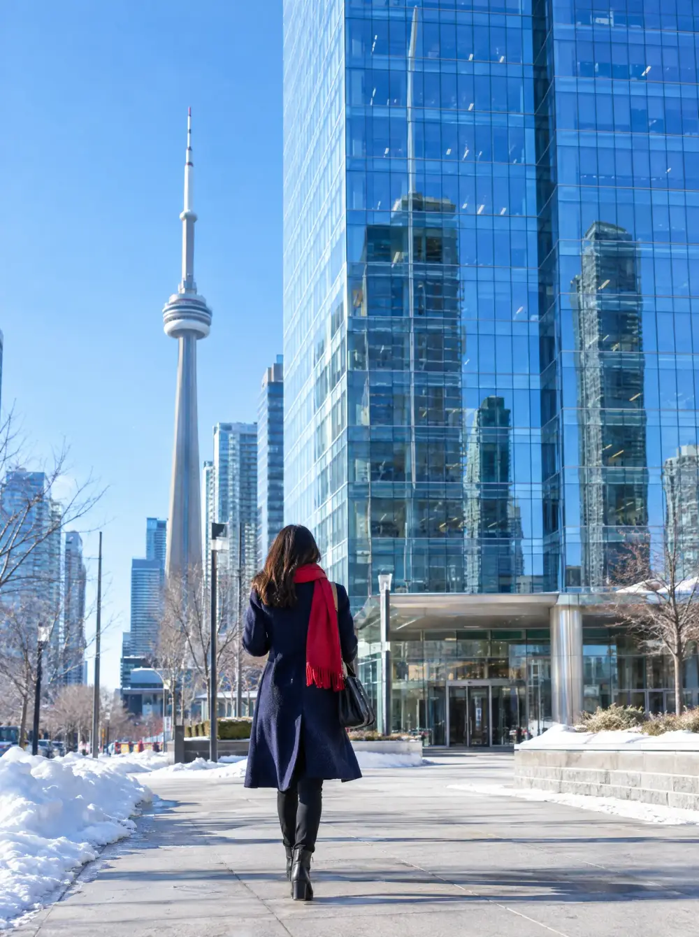 A person in a red scarf walking toward a glass office tower in downtown Toronto on a sunny winter day