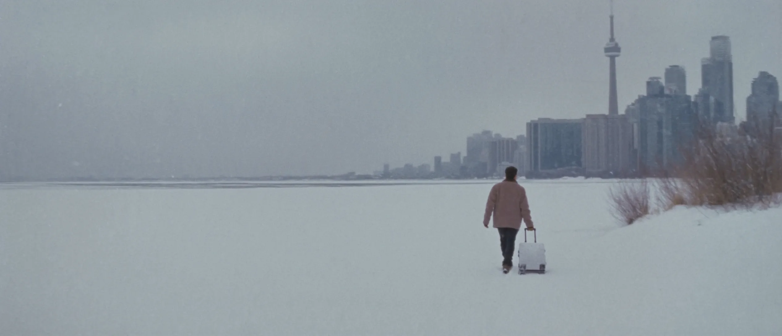 A traveller with a suitcase walks along the snowy shore of Lake Ontario toward the Toronto skyline