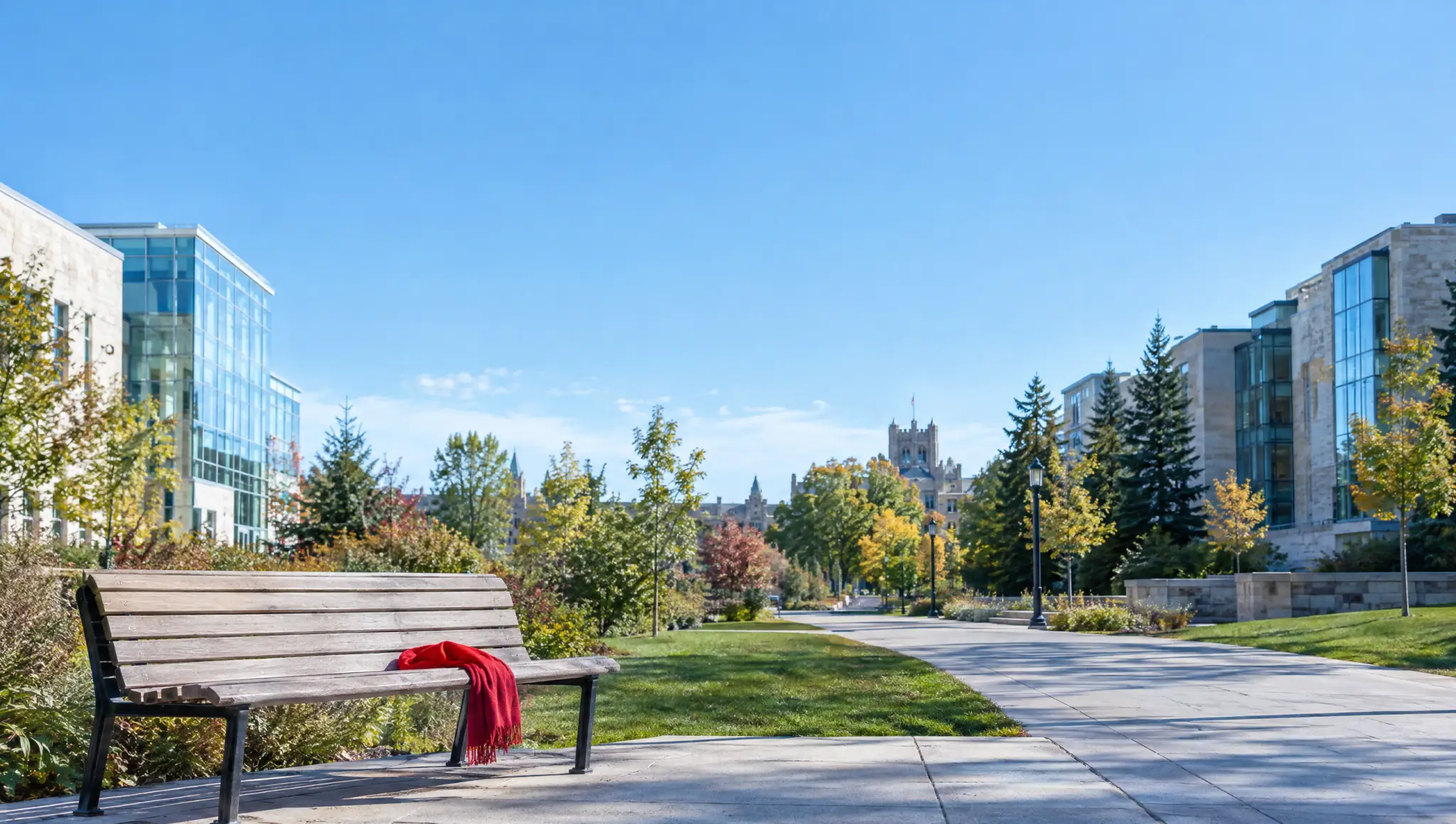 A sunny Canadian university walkway between glass and stone buildings, with a red scarf left on an empty bench